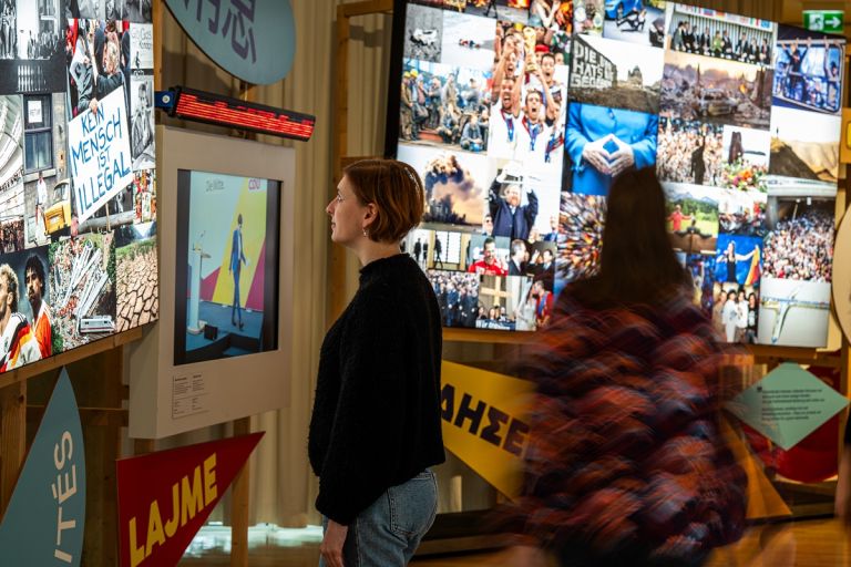 Zwei Personen stehen in Raum mit Bildschirmen und Nachrichtenbilderflut an den W&auml;nden und schauen diese an